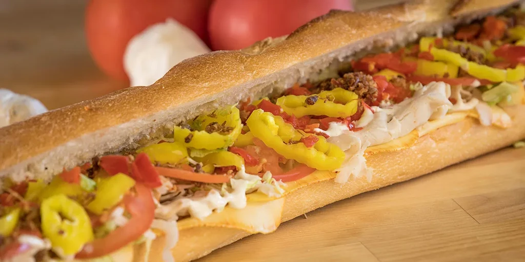 Hoagie with deli meat, cheese, lettuce, tomato, banana peppers on wood.