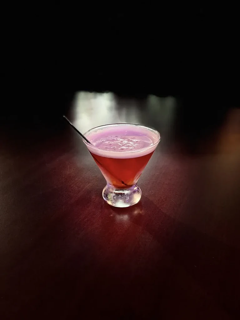 Red cocktail in a martini glass, frothy top, dark straw.