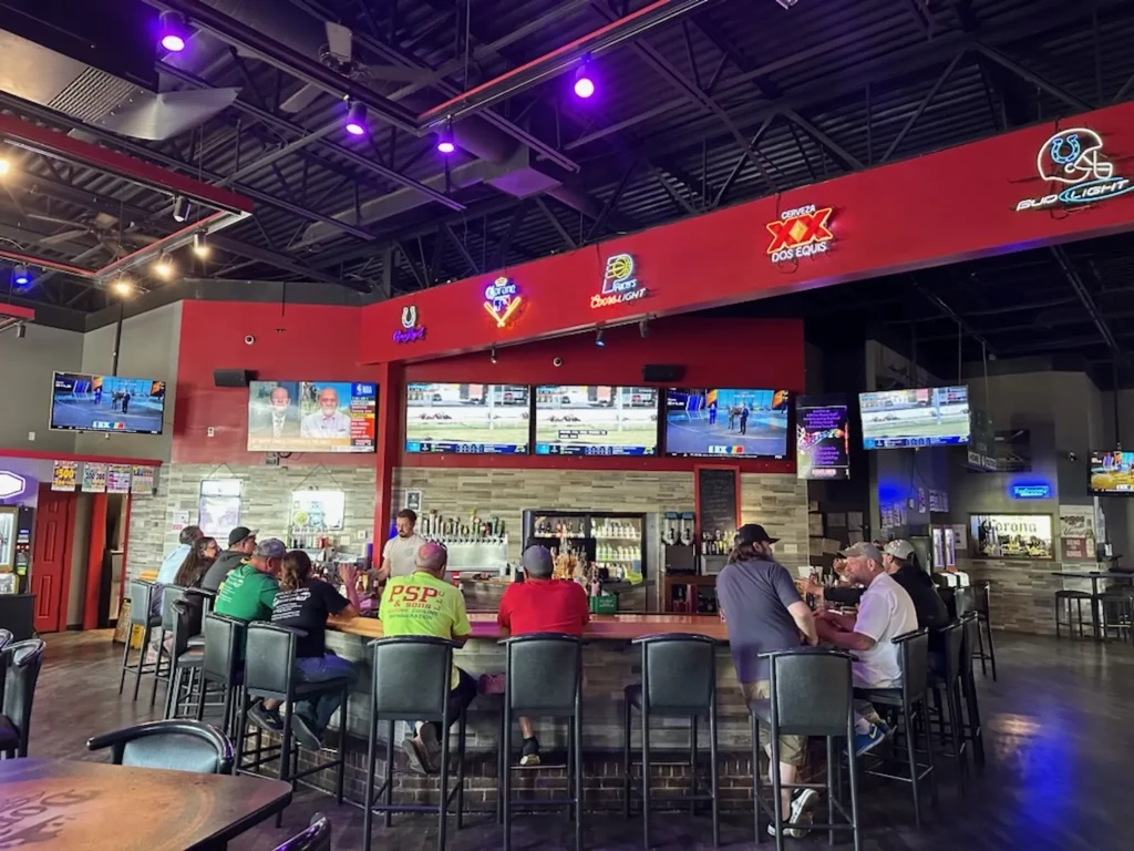 Sports bar interior with U-shaped bar, patrons, TVs, and neon signs.