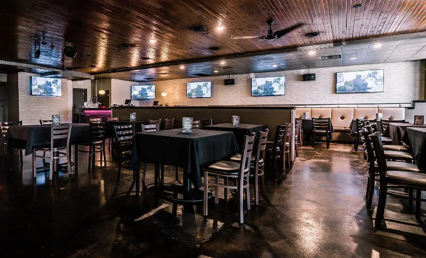 Restaurant interior with dark tables, booth seating, purple bar, and TVs.