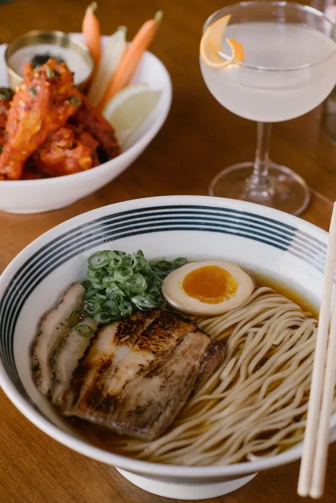 Restaurant table close-up: steaming ramen, chicken wings, and a cocktail.