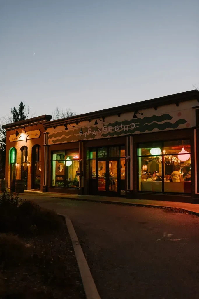 STRANGEBIRD restaurant exterior at twilight, with vibrant mural and inviting interior lights.