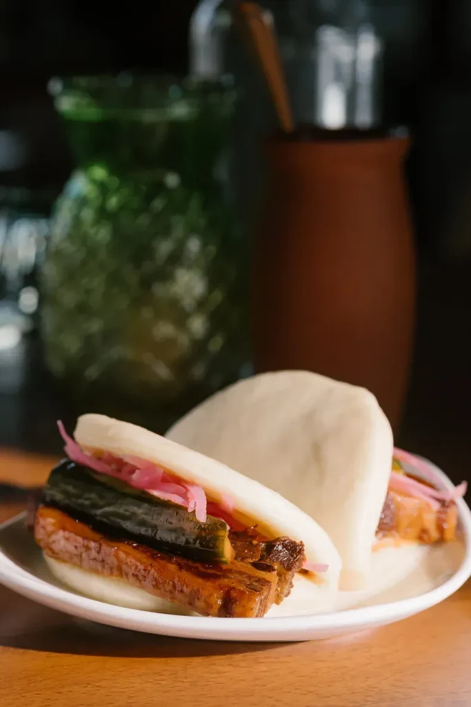 Two bao buns with pork belly, pickled cucumber, red onions on white plate.