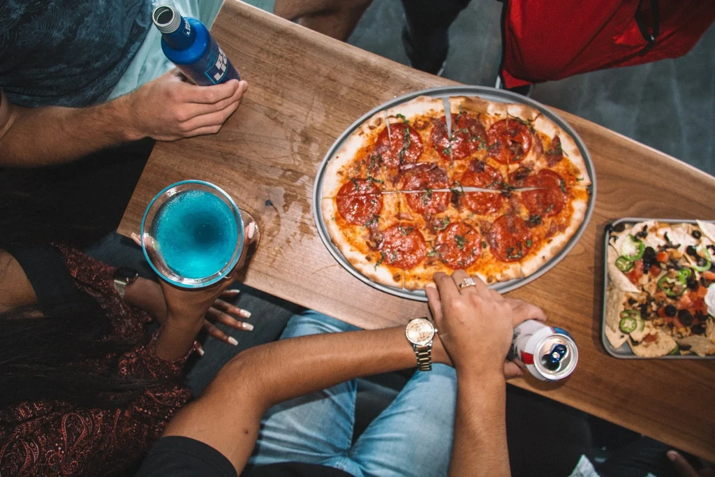 Overhead: Hands share pepperoni pizza, nachos, beer, and a blue cocktail.