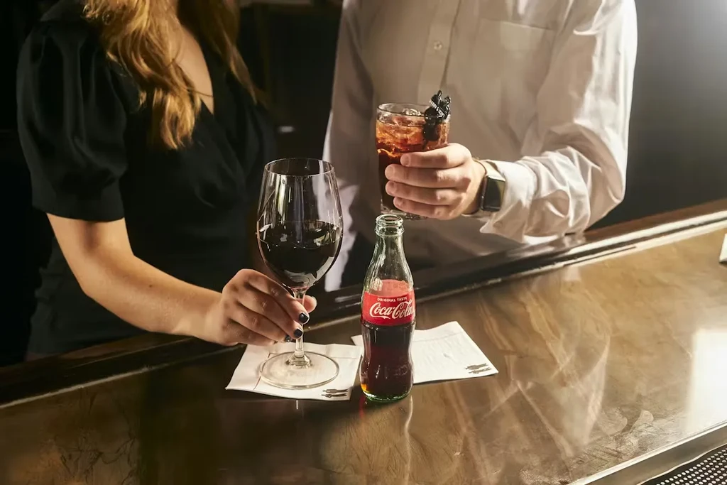 Hands holding red wine, a cocktail, and Coca-Cola on a glossy bar.
