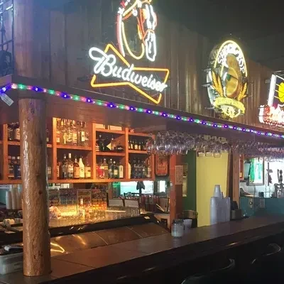 Dark wooden bar with Budweiser neon sign, liquor, and hanging glasses.