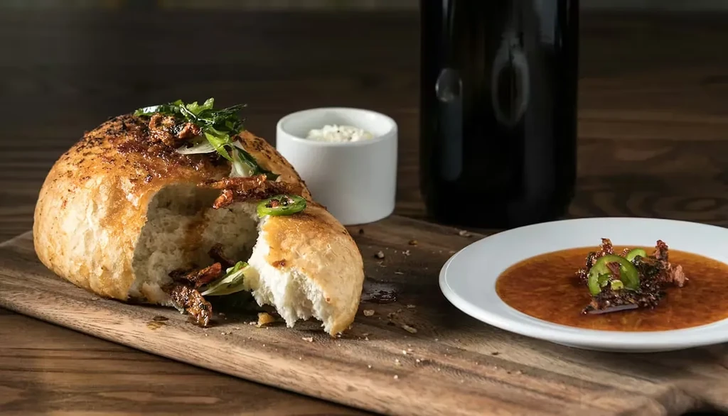 Torn bread with meat, herbs, cheese, liquid, and bottle on wood.