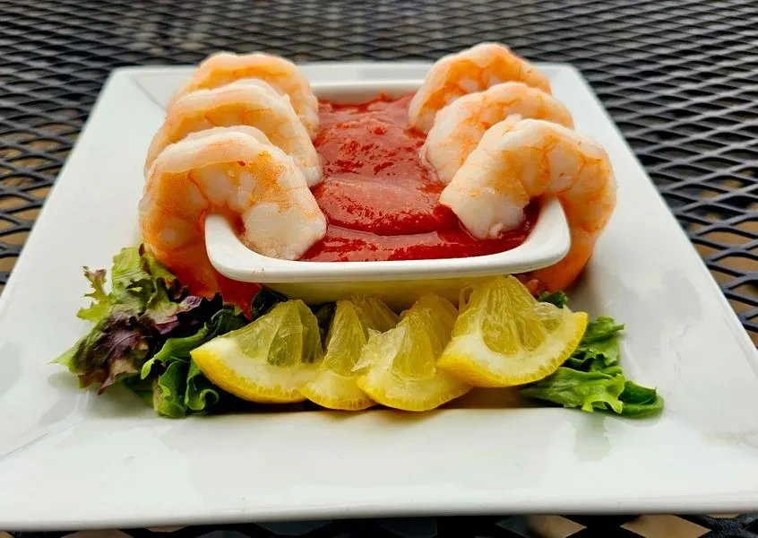 Eight cooked shrimp around red cocktail sauce with lemon and lettuce.