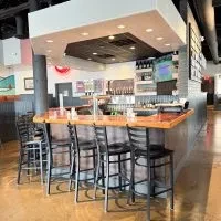 Well-lit modern bar, L-shaped wood counter with black stools.