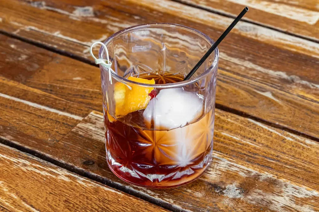 Layered cocktail in cut-glass with spherical ice, orange peel, straw.