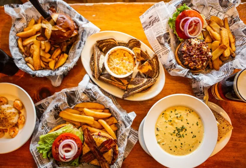 Wooden table with burgers, fries, dip, bread, beer, and soup.