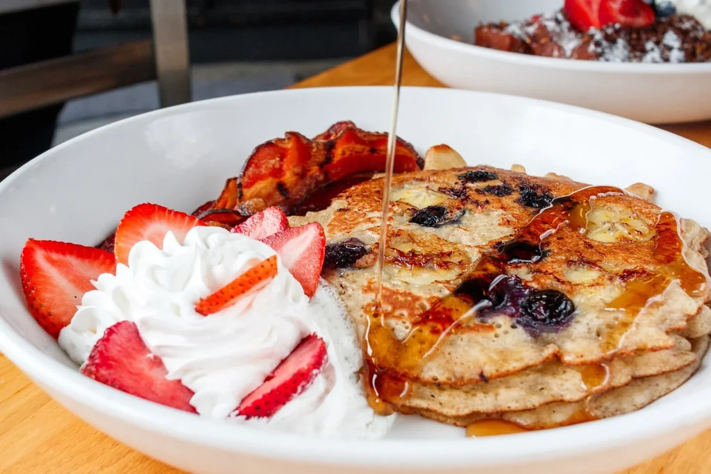 Fluffy blueberry pancakes with syrup, strawberries, whipped cream, and bacon.