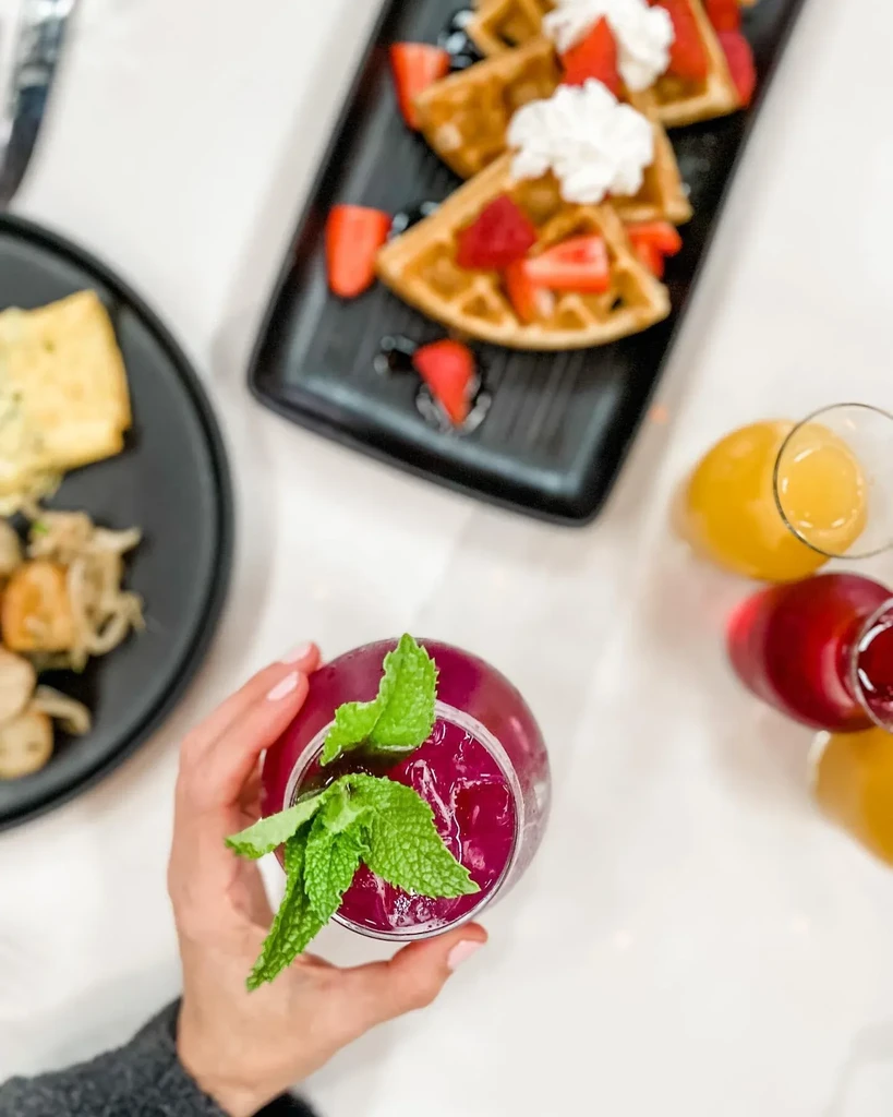 Hand holds purple drink; waffles with fruit and savory brunch dishes.
