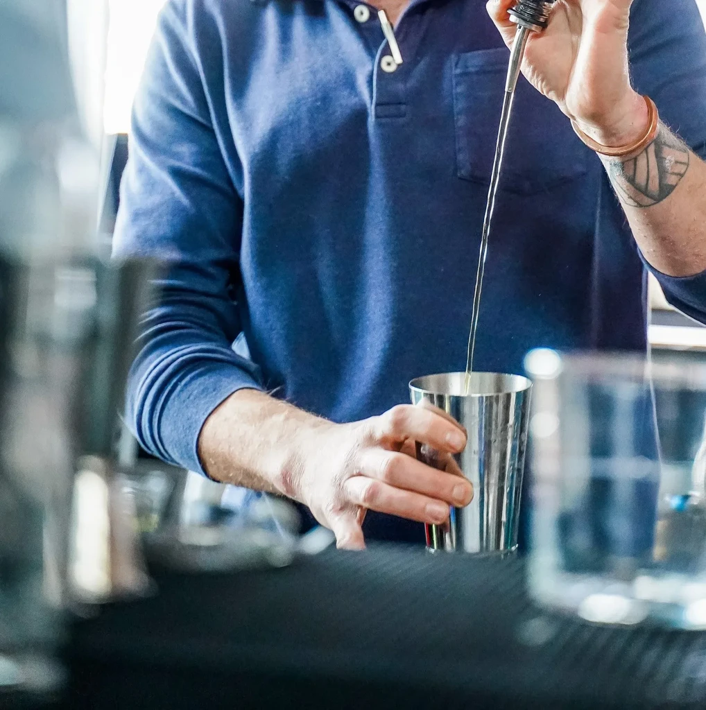 Bartender in navy polo pouring liquid into a cocktail shaker.