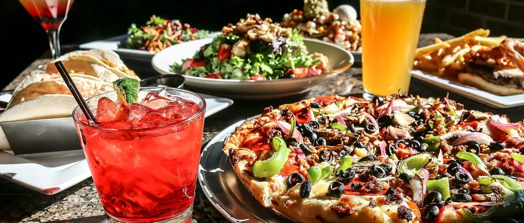 Vibrant table with loaded pizza, red cocktail, tacos, salads, and drinks.