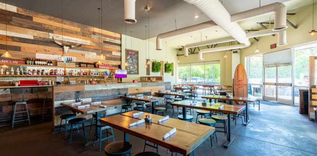 Empty rustic-industrial restaurant with wood, exposed ducts, bull skull, and surfboard.