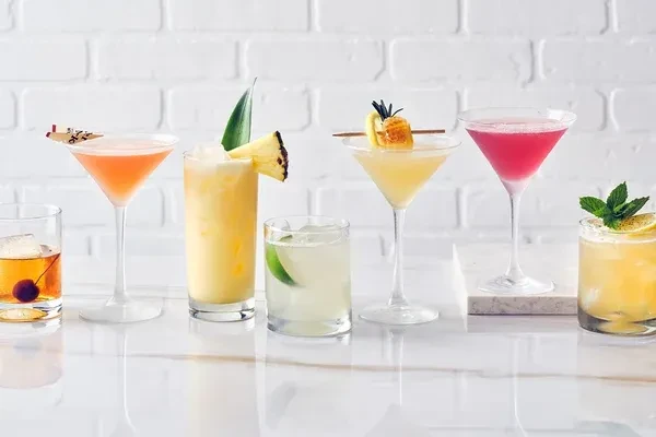 Line of six vibrant cocktails on a white reflective surface.