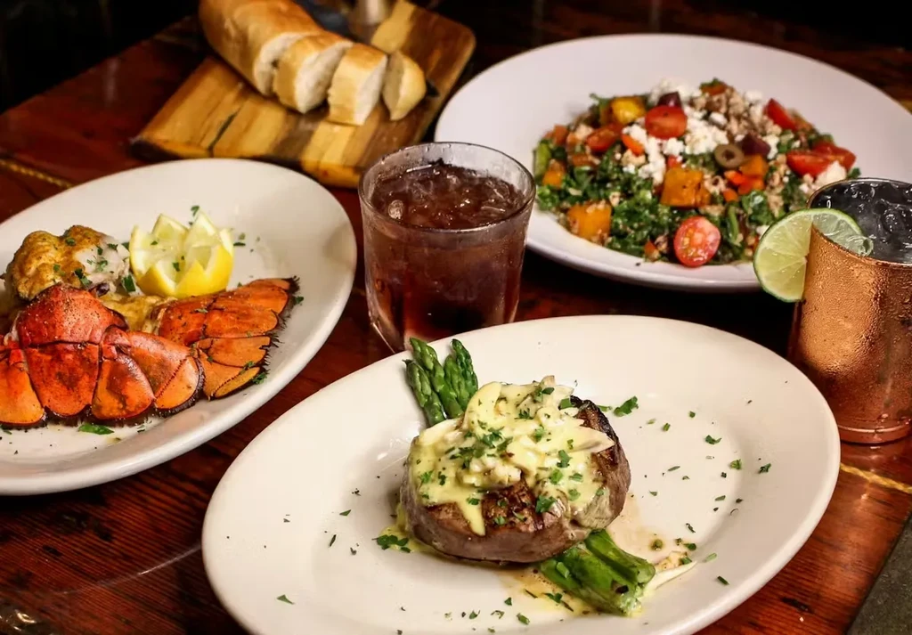 Restaurant table with seared steak, lobster, salad, and two drinks.