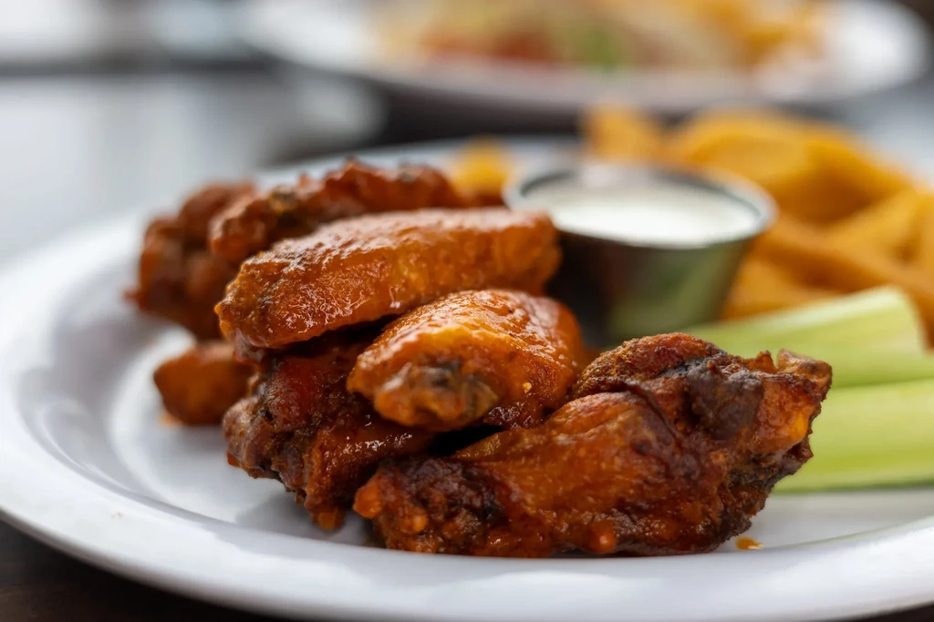 Plate of glossy buffalo wings, celery, dipping sauce, and blurred fries.