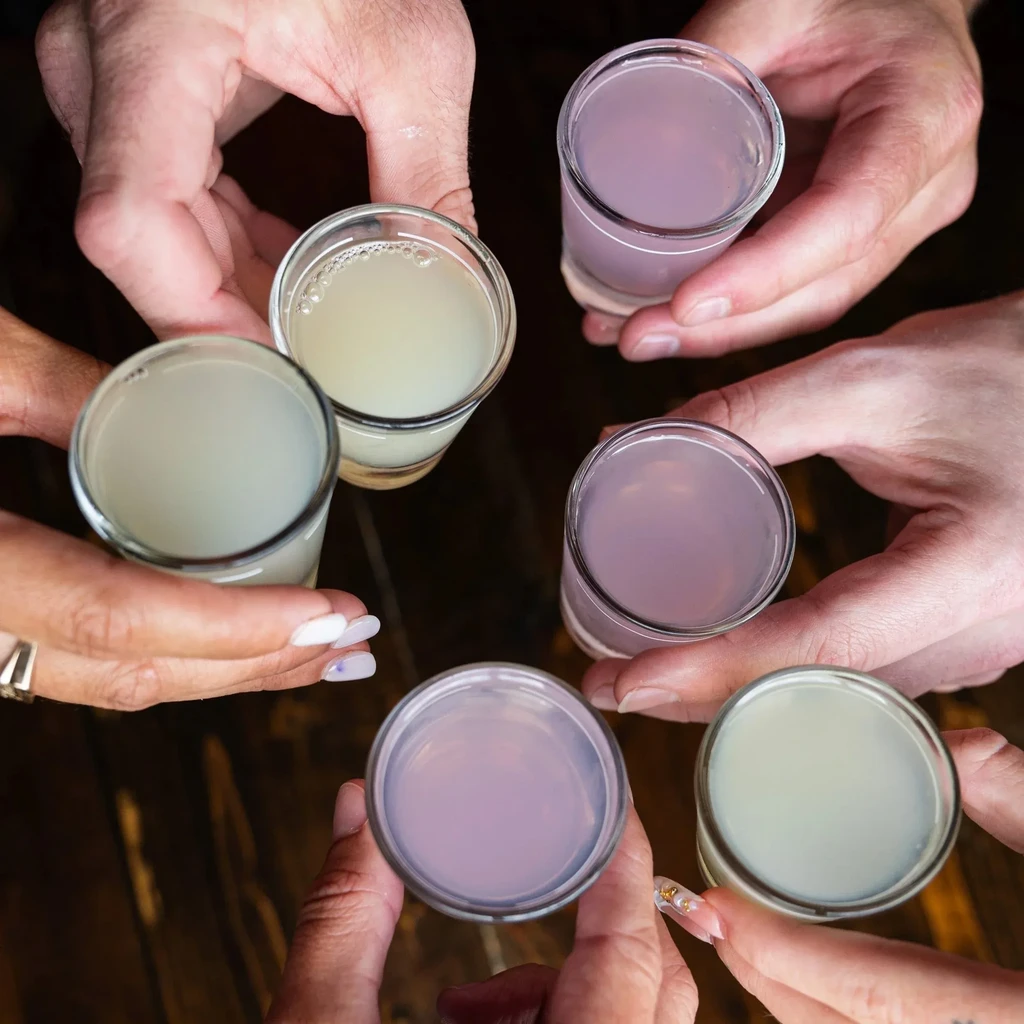 Diverse hands clink six shot glasses of yellow and purple liquids.