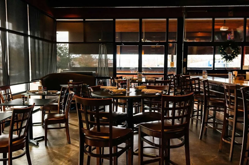 Empty modern restaurant interior with dark wood tables, bar, and holiday wreath.