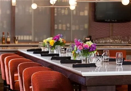 Elegant bar interior with communal table, vibrant flowers, and orange chairs.