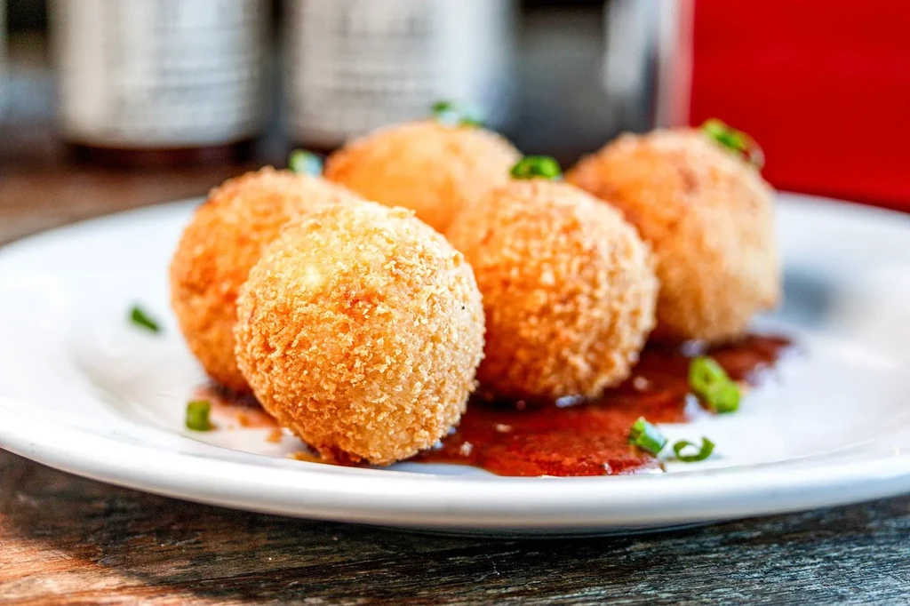 Five golden-brown breaded spheres on a plate with sauce and chives.