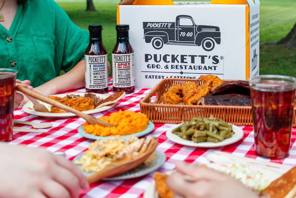 Puckett's outdoor picnic with BBQ, sides, and sauces on checkered tablecloth.