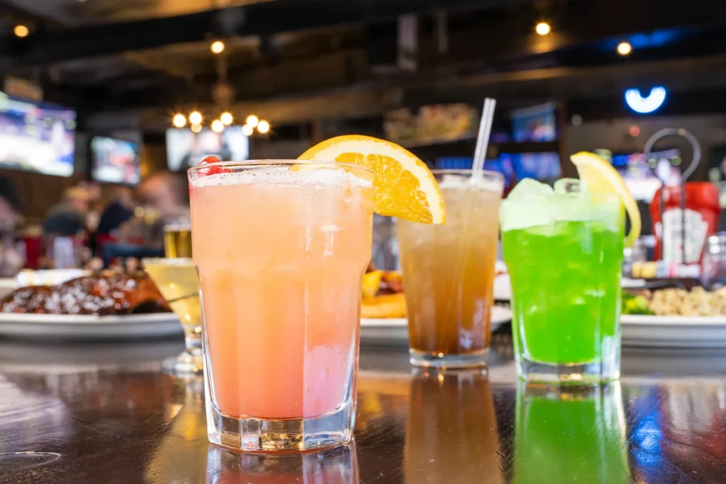 Colorful cocktails on a reflective bar top in a bustling sports bar.