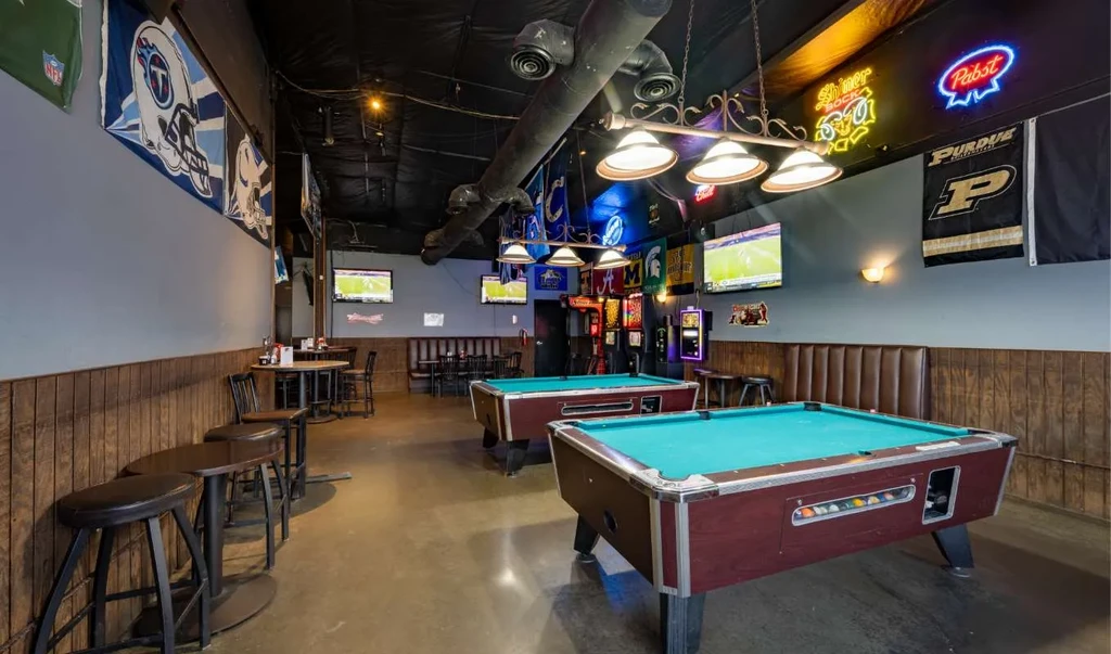 Two blue felt pool tables in a sports bar with TVs and flags.