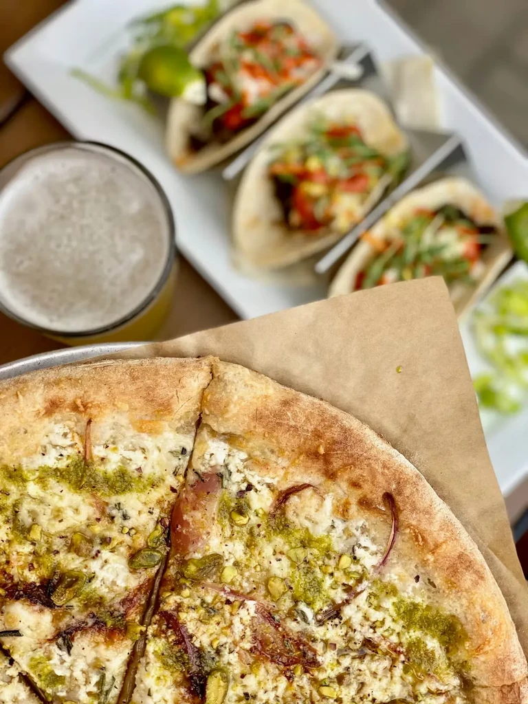 Overhead: pizza with cheese, pesto, onions, nuts; beer; blurred tacos.