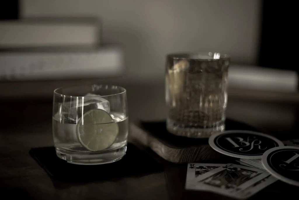 Two glasses, one with lime, playing cards, and coasters on dark wood.