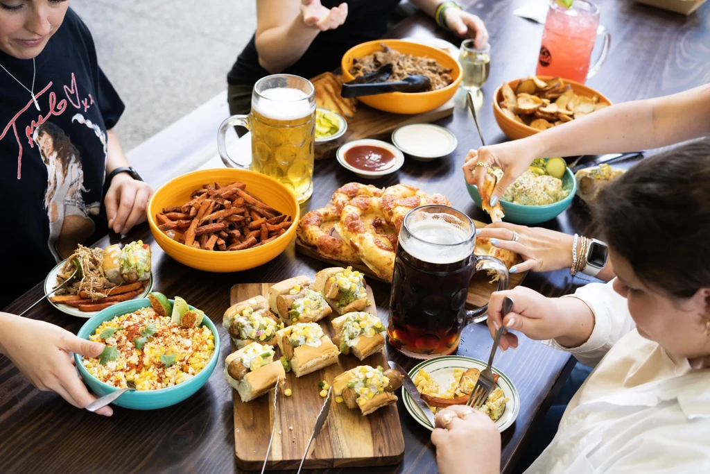 Hands reaching for diverse food and drinks on a wooden table.