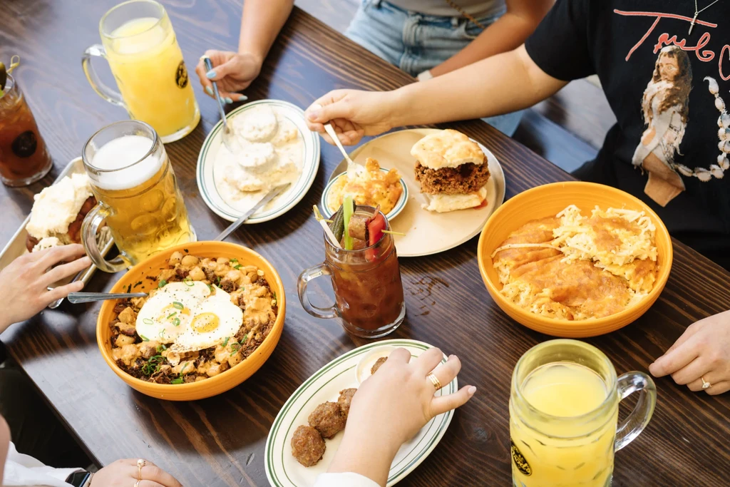 Various brunch dishes, drinks, and hands reaching on a dark dining table.