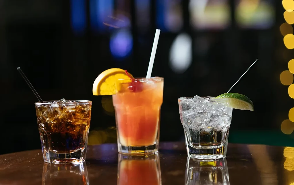 Three colorful cocktails with ice and garnishes on a reflective surface.