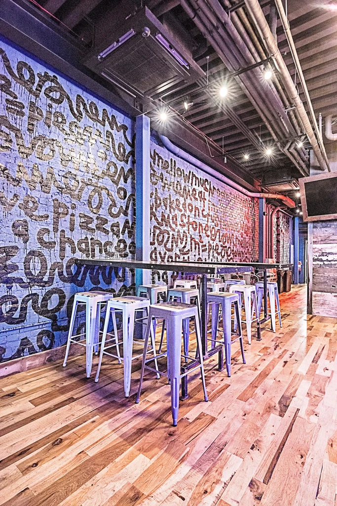 Industrial bar: high-top table, graffiti brick walls, exposed pipes, bright lights.