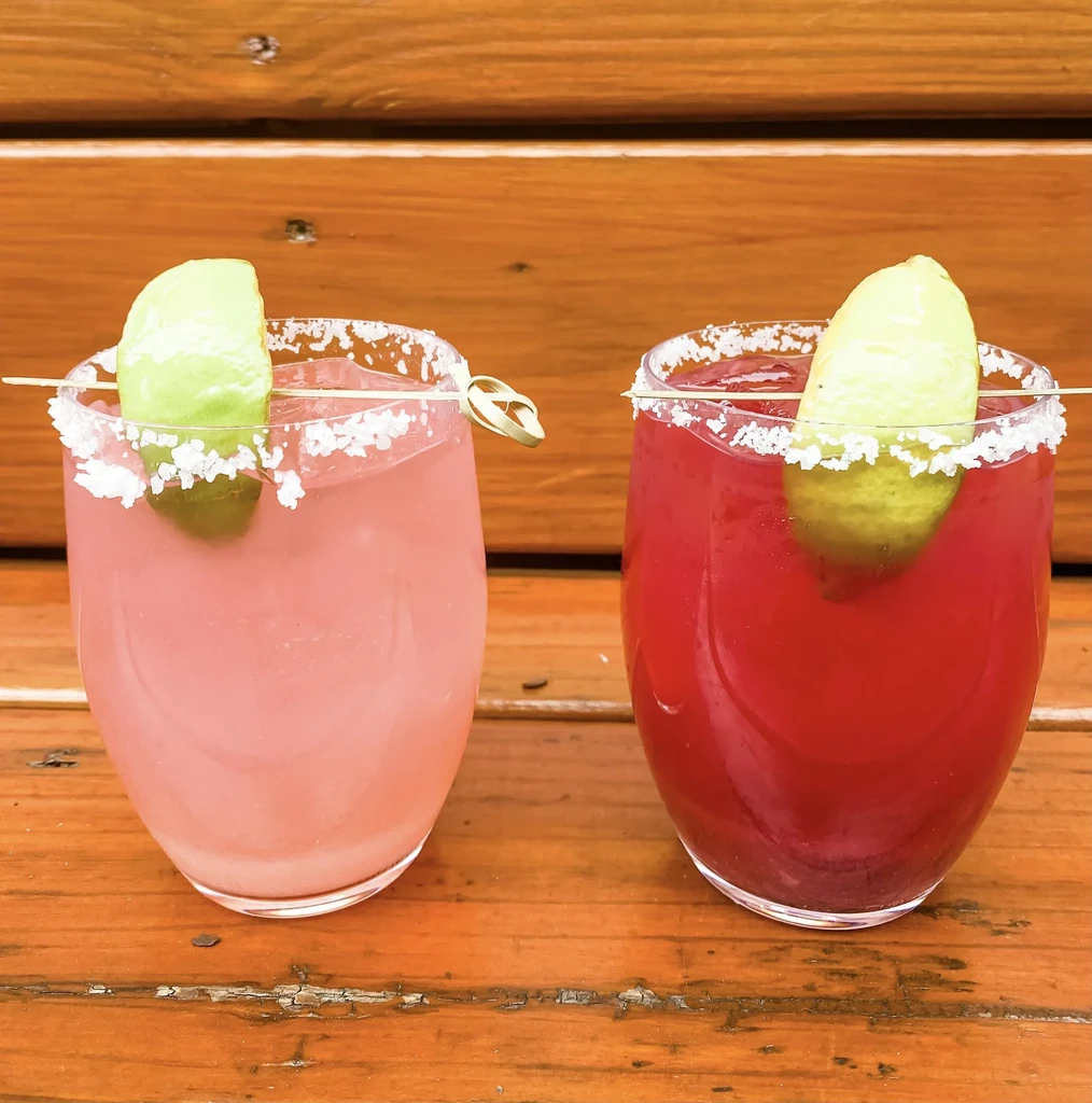 Pink and red salt-rimmed cocktails with lime on rustic wood.