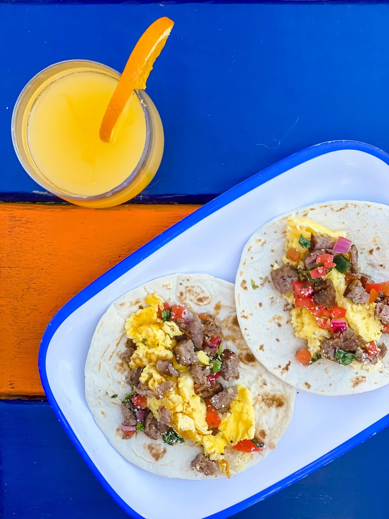 Top-down view of two breakfast tacos, orange juice, vibrant background.