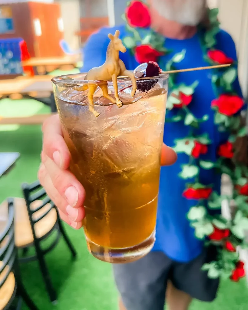 Hand holds an amber cocktail with a toy horse and cherry garnish.