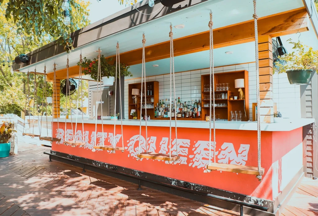 Outdoor bar with "REALITY QUEEN" coral-orange front and swing seats.