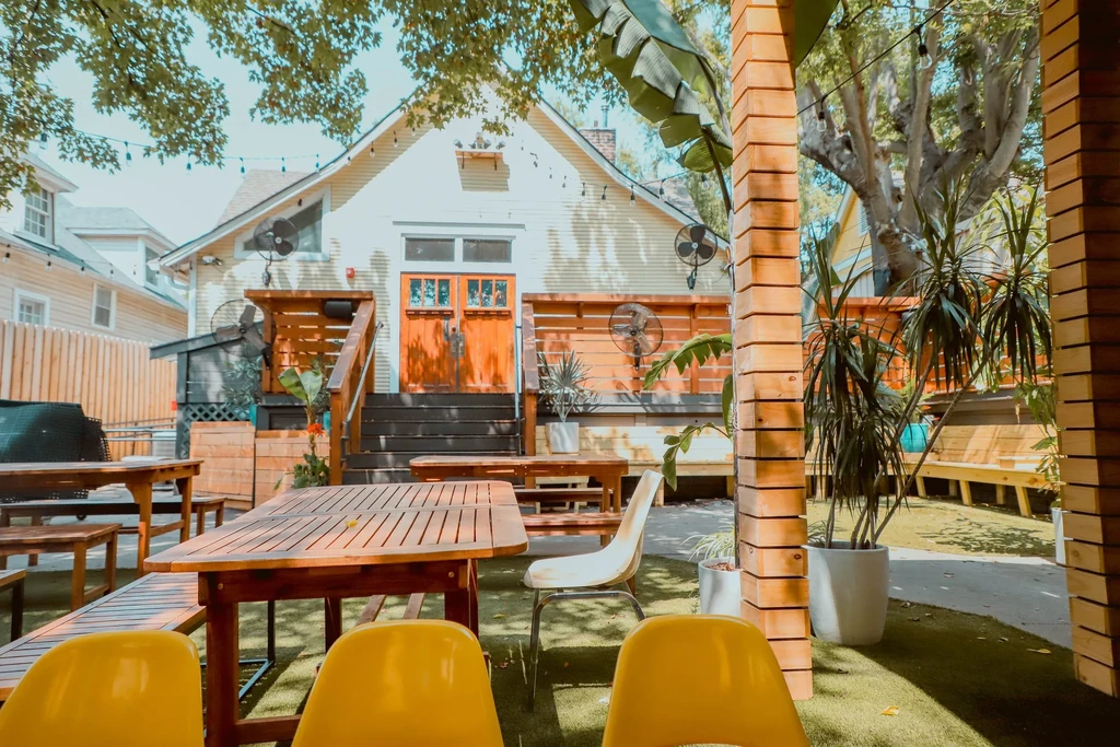 Lush restaurant patio with wooden tables, yellow chairs, string lights.