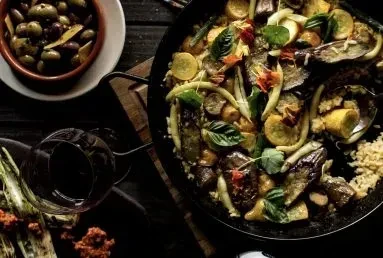 Rustic table with paella, olives, red wine, and grilled vegetables.