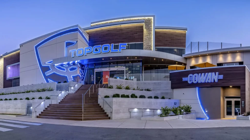 Modern Topgolf complex exterior at twilight, vibrant blue neon and seating.