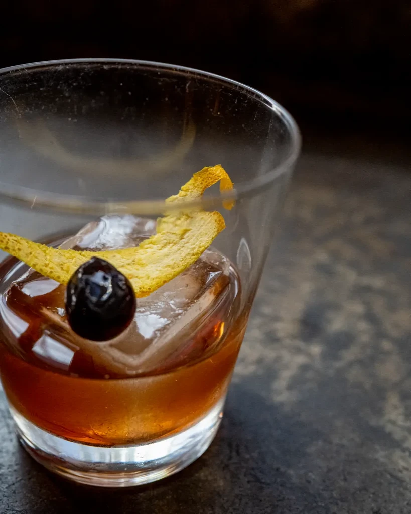 Amber cocktail with large ice, dark cherry, and orange peel twist.