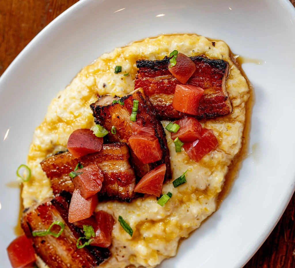 Creamy grits with crispy pork belly, diced red cubes, and chives in a white bowl.