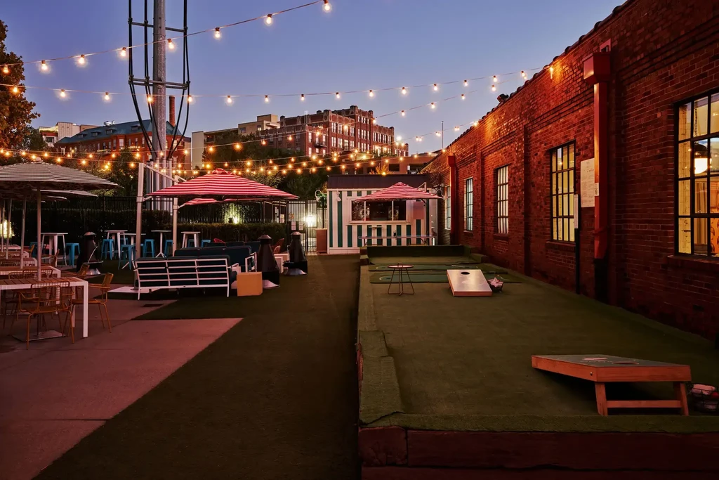 Outdoor evening patio with dining, mini-golf, and cornhole under string lights.