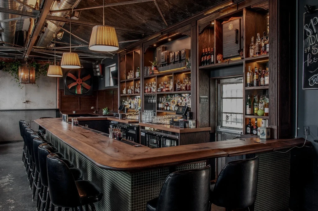 Industrial bar with long wooden bar, mosaic tiles, illuminated bottle display.
