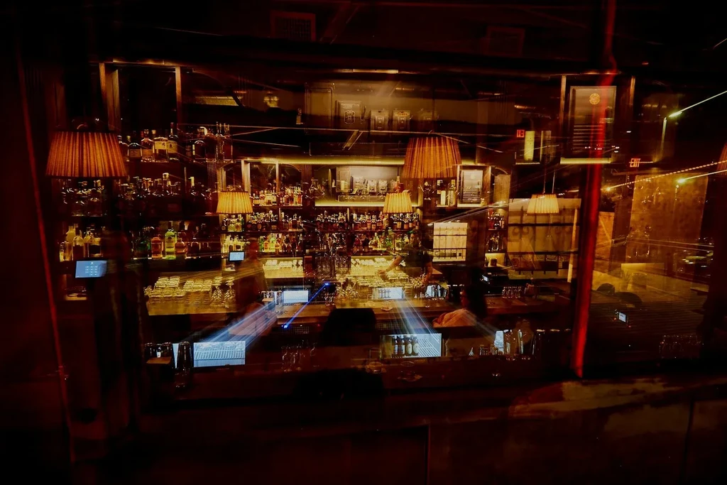 Dimly lit, well-stocked bar with warm lights and blurred figures.