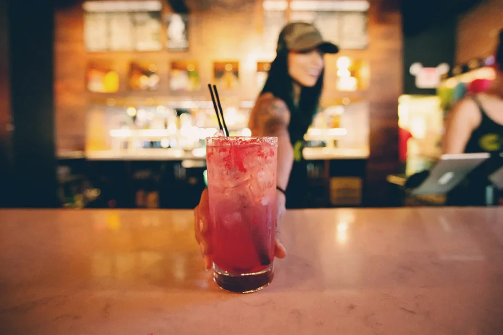 Vibrant red-pink cocktail, sugared rim, blurred bartender presenting at bar.