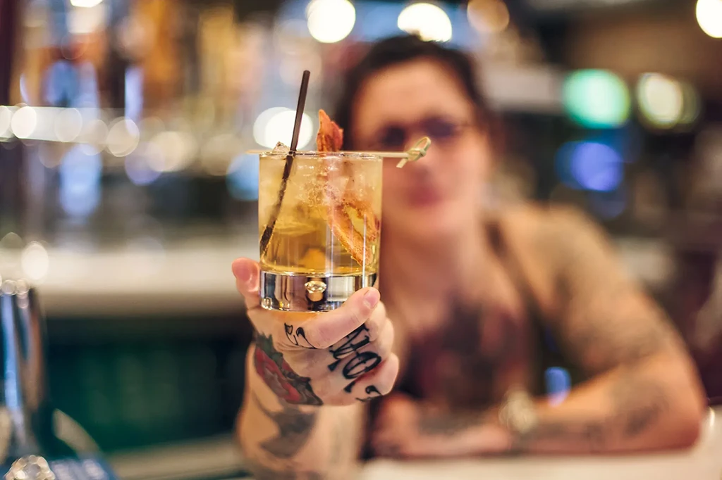 Close-up: Tattooed hand holds amber cocktail with garnish, blurred background.